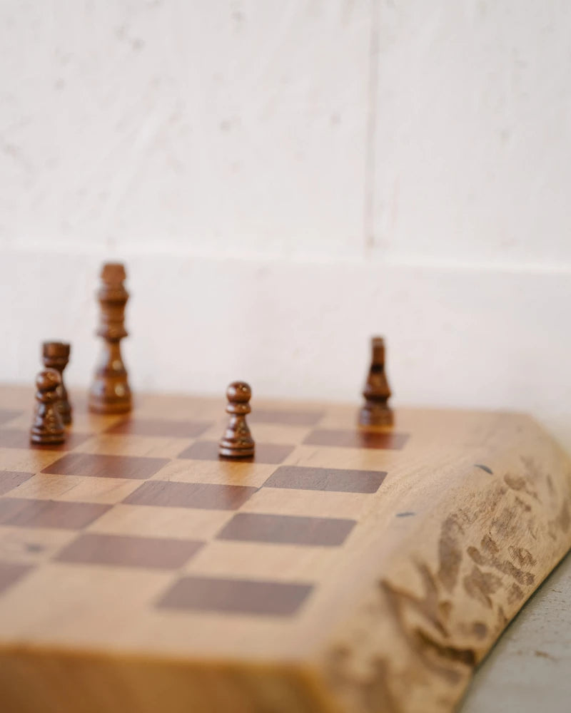 Wooden chessboard with four dark wooden pieces on a light background. Pieces include a king, two pawns, and a bishop. Edges of the chessboard are rough and natural.