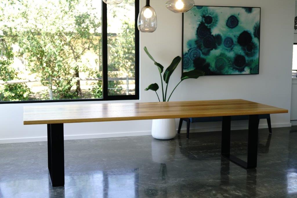 A Timber Grooves American White Oak Dining Table with a black steel table base stands in a room with a large window, a white potted plant, and a colorful abstract painting on the wall. The floor is polished concrete.
