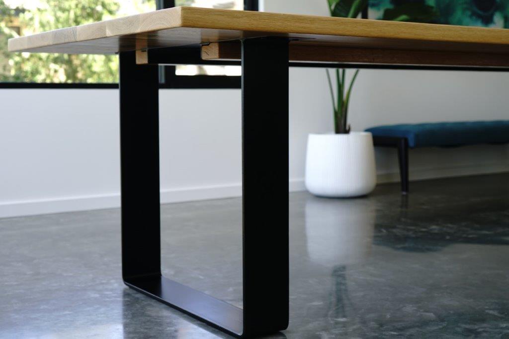 Close-up of a modern Timber Grooves American White Oak Dining Table with a wooden top and black steel table base. A white planter with a plant and a blue bench are visible in the background, showcasing this piece of handcrafted dining room furniture.