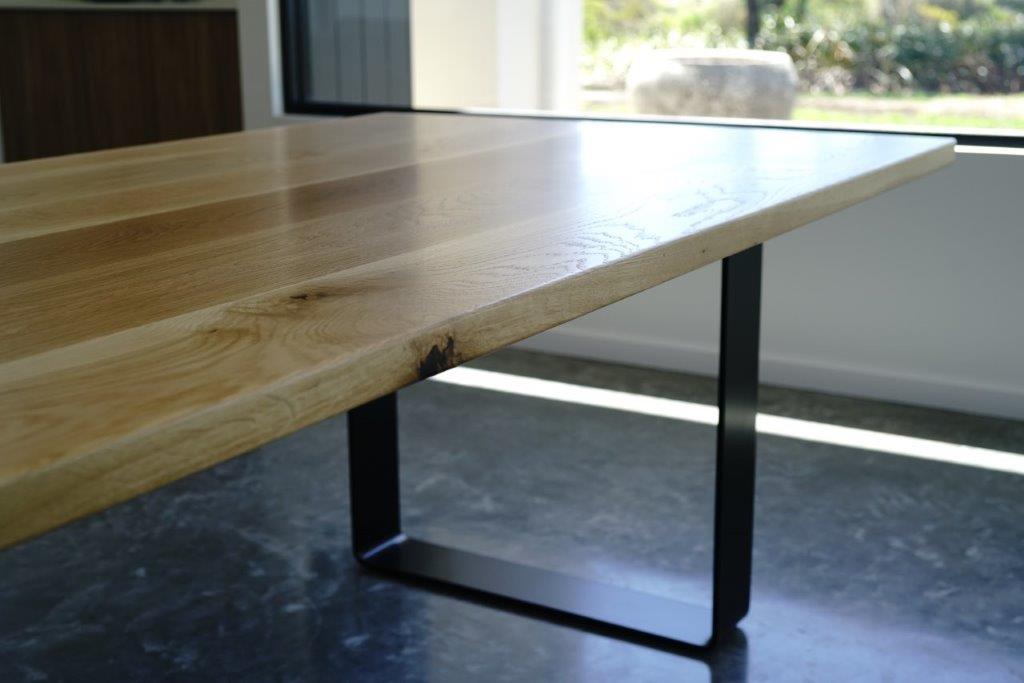 A close-up view of a Timber Grooves American White Oak Dining Table with a black steel table base set on a dark stone floor near a window with natural light coming in.