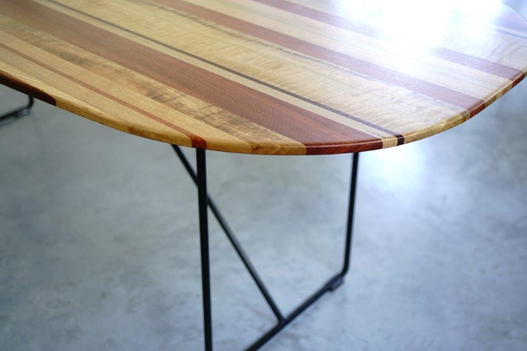A handcrafted **Timber Grooves Surfboard Dining Table** with a glossy surface and metal legs. The tabletop, inspired by surfboard aesthetics, features a striped pattern made of different wood types, showcasing unique grain patterns.