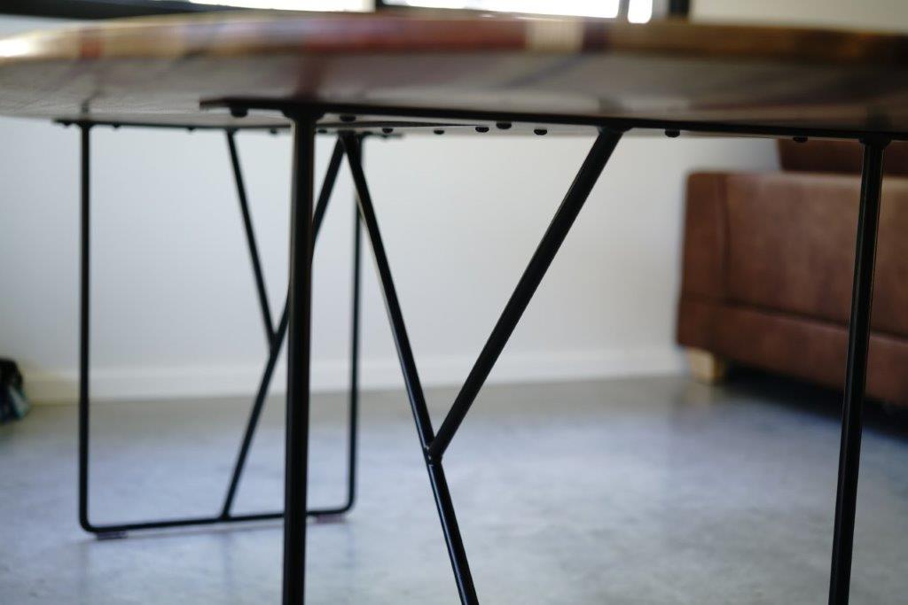 Close-up of the metal legs supporting a Timber Grooves Surfboard Dining Table with unique grain patterns on a smooth concrete floor, with part of a brown sofa visible in the background.