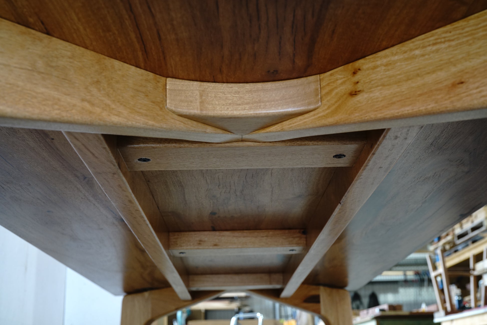 Close-up view of the underside of a luxury handcrafted wooden table, revealing intricate support structures and joinery details in a workshop setting. This Marri Dining Table from Timber Grooves exemplifies exquisite craftsmanship.