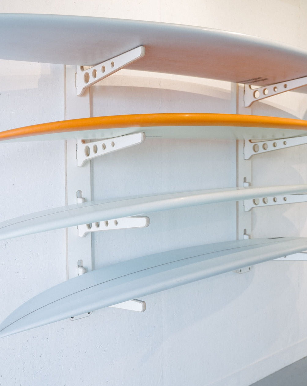Three surfboards are neatly arranged on the Timber Grooves Surfboard Rack Wall Mount. The top surfboard is orange, and the two below it are white.