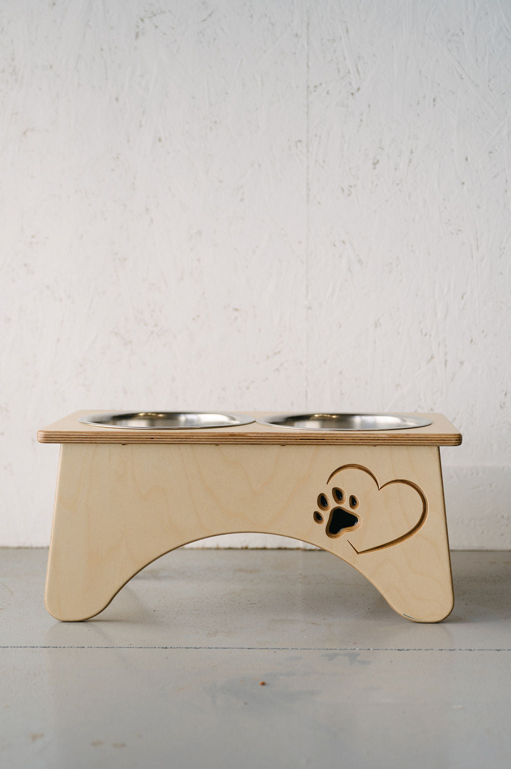 The Timber Grooves Double Raised Dog Bowl Stand features a wooden base with two metal bowls. Designed to reduce symptoms of bloat and alleviate skeletal and joint pain, one side showcases a charming heart cutout with a paw print inside. Perfect for your pet's health and comfort.