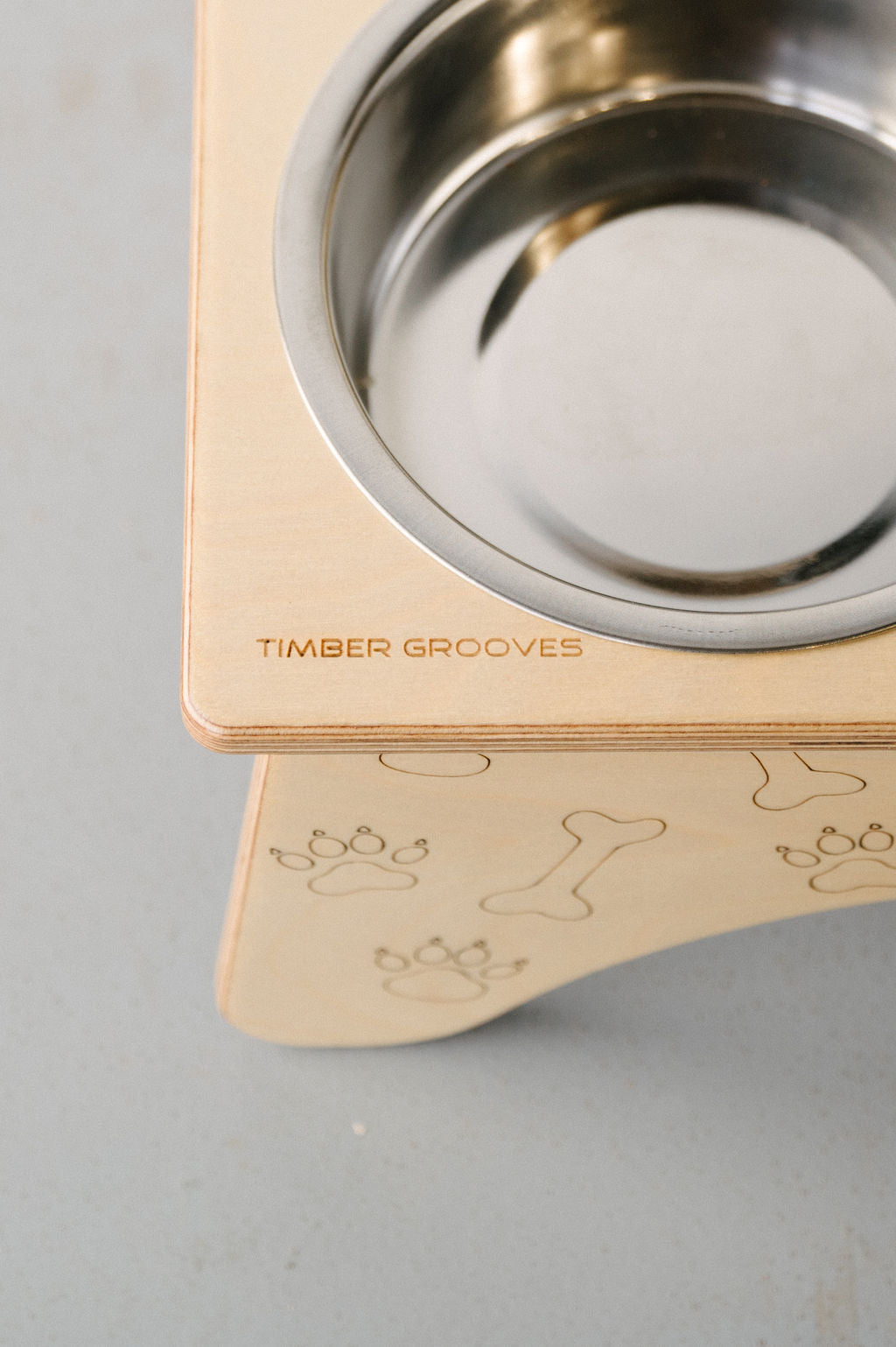 A wooden pet feeding station with engraved patterns of bones and paw prints, holding a round stainless steel bowl. The brand name "Timber Grooves" is etched into the wooden surface. This Double Raised Dog Bowl Stand helps reduce symptoms of bloat and supports pets with skeletal and joint pain.