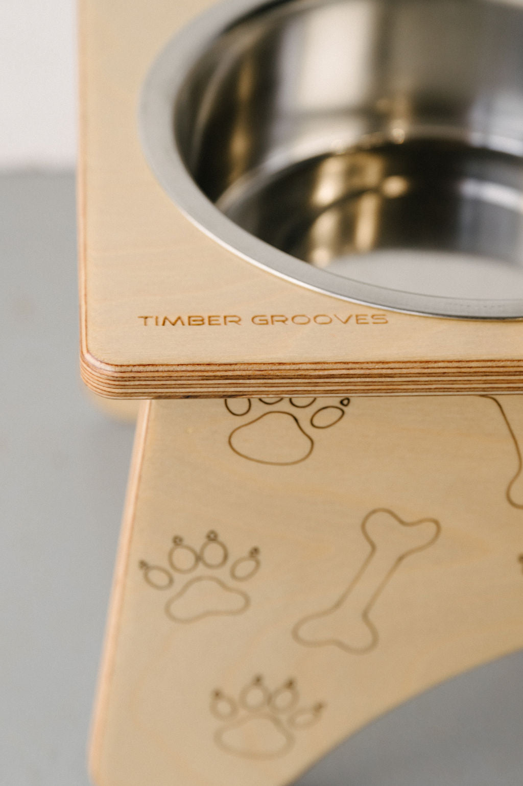 Close-up of a wooden Double Raised Dog Bowl Stand with stainless steel bowls, featuring carved paw prints, a bone design, and the brand name "Timber Grooves" engraved on the side. This elevated feeder helps reduce symptoms of bloat, promoting better digestion for your furry friends.