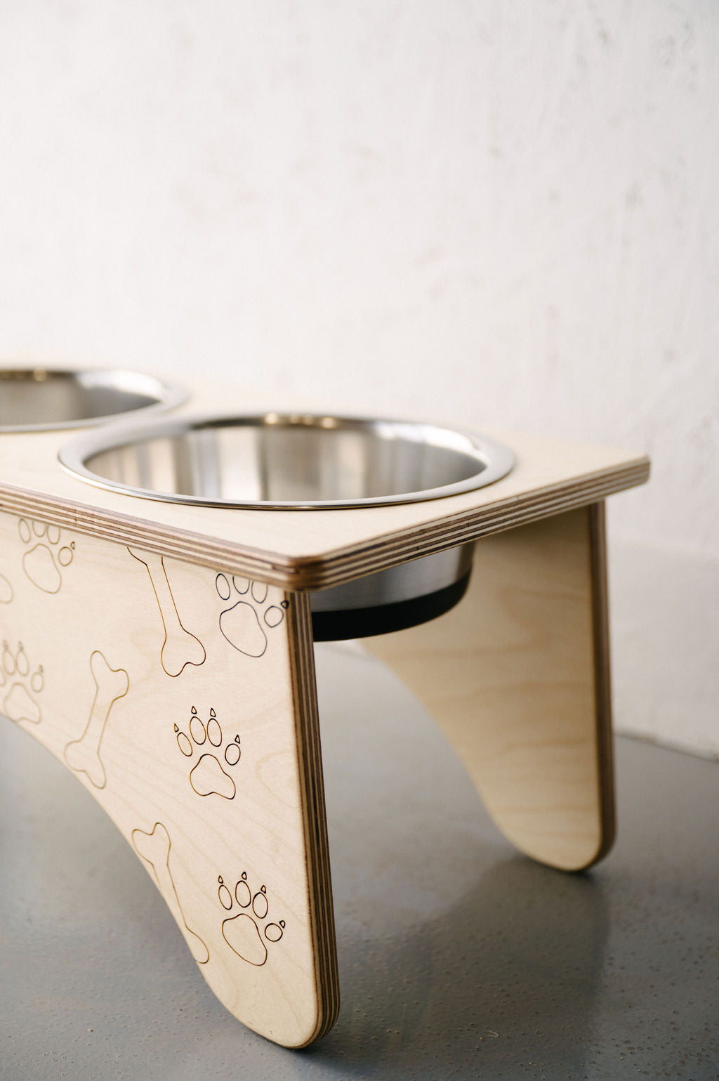 A Double Raised Dog Bowl Stand by Timber Grooves made of wood with two stainless steel bowls, featuring engraved paw and bone designs on its side. This elevated feeder helps reduce symptoms of bloat while alleviating skeletal and joint pain in pets.