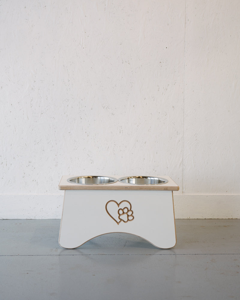 A Double Raised Dog Bowl Stand by Timber Grooves with stainless steel bowls and a heart with a paw print design on the front helps reduce symptoms of bloat and alleviate skeletal and joint pain. This white elevated pet bowl stand is placed against a plain white wall and gray floor.