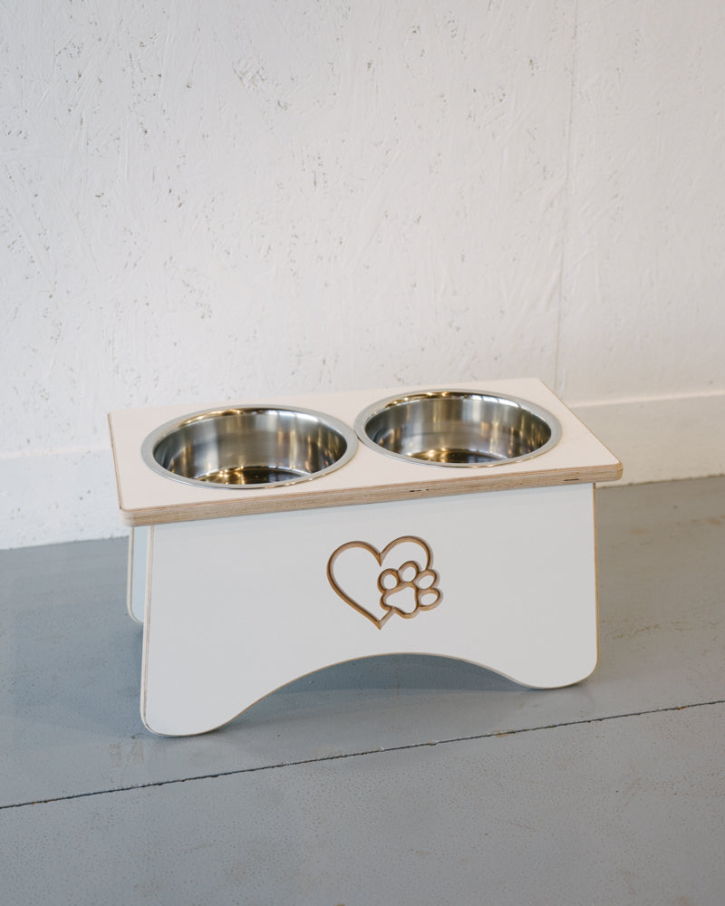 The Timber Grooves Double Raised Dog Bowl Stand in white, featuring two stainless steel bowls and a heart with a paw print design on the front, is perfect for reducing symptoms of bloat and alleviating skeletal and joint pain in pets.
