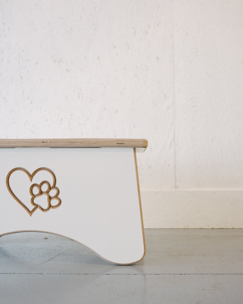 Side view of a small white wooden stool with a carved heart and paw print design, placed on a light grey floor against a plain white wall. This charming piece could complement any decor while also being the perfect companion to Timber Grooves Double Raised Dog Bowl Stand that helps reduce symptoms of bloat in pets.