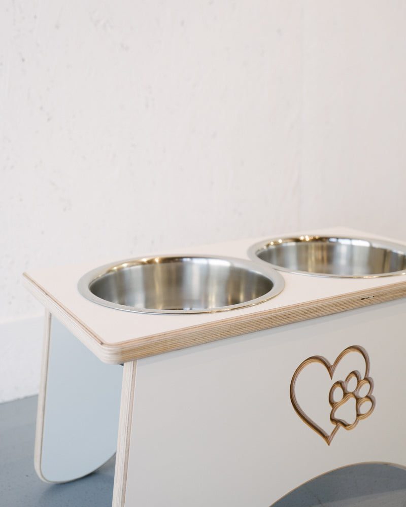 A white wooden Timber Grooves Double Raised Dog Bowl Stand with two stainless steel bowls and a heart with paw print design on the front stands against a plain white wall, designed to reduce symptoms of bloat and alleviate skeletal and joint pain in pets.