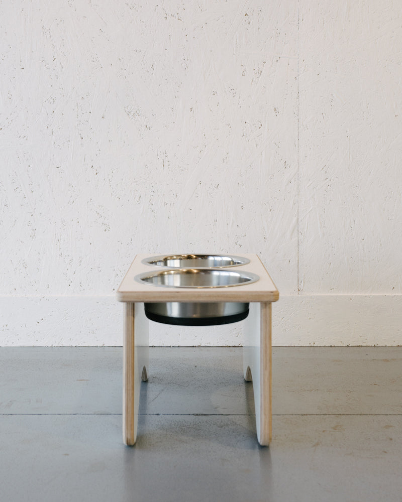 A Timber Grooves Double Raised Dog Bowl Stand with two stainless steel bowls is placed on a gray floor against a white textured wall—perfect for reducing symptoms of bloat and easing skeletal and joint pain.