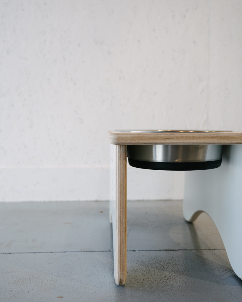 A sleek Timber Grooves Double Raised Dog Bowl Stand with a wooden base and stainless steel bowls is placed on a polished concrete floor against a white wall, designed to reduce symptoms of bloat and alleviate skeletal and joint pain in your furry friend.