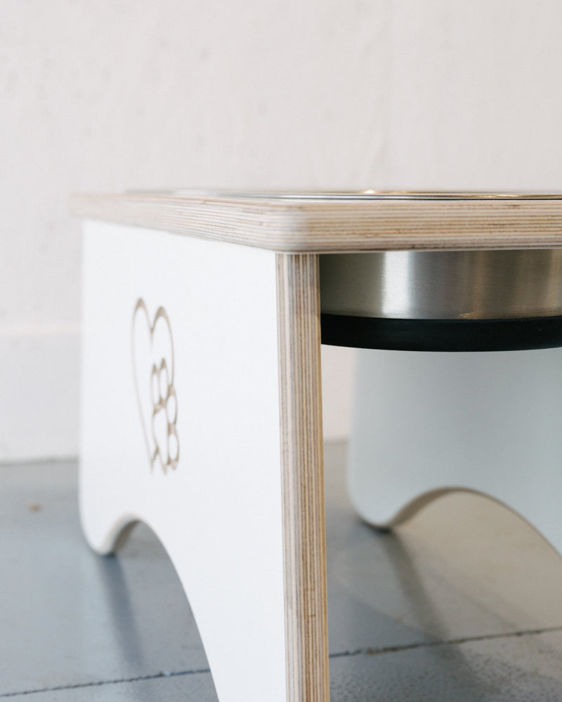 Close-up of a Timber Grooves Double Raised Dog Bowl Stand, a minimalist wooden pet feeder with heart and paw print design, featuring stainless steel bowls on a light gray floor. Ideal for reducing symptoms of bloat and minimizing skeletal and joint pain in pets.