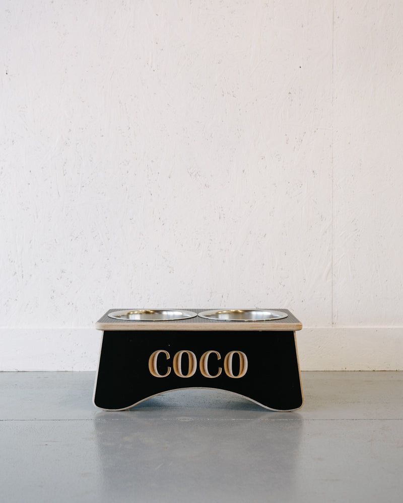 A black Timber Grooves Double Raised Dog Bowl Stand with two stainless steel bowls and the name "COCO" on the front, placed on a grey floor against a white wall. Perfect for reducing symptoms of bloat and aiding in skeletal and joint pain relief.