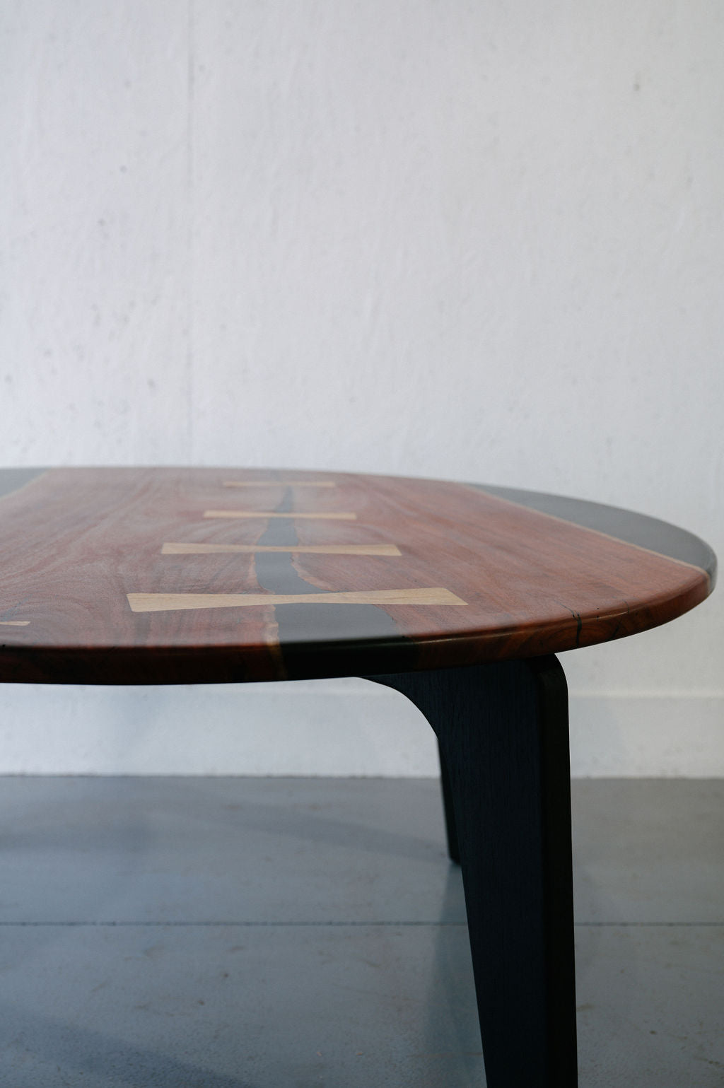 Close-up of a Timber Grooves Jarrah Tuxedo Coffee Table with black legs, featuring an inlaid pattern of light wood and marri bowties running along the length of the tabletop. The background is a plain white wall and gray floor.