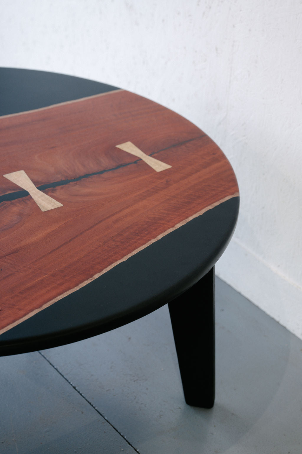 Close-up of a Timber Grooves Jarrah Tuxedo Coffee Table with a dark border, showcasing a unique wood grain pattern with light-colored inlays. Black resin marri bowties accent the design. The table is placed against a white textured wall on a gray floor.