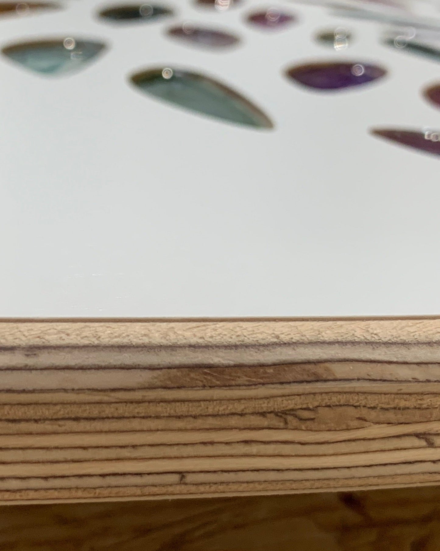 A close-up of colorful, teardrop-shaped inlays embedded in a light wooden surface, resembling Resin leaves Tree of Life in White by Timber Grooves, with part of the edge showing visible wood grain.