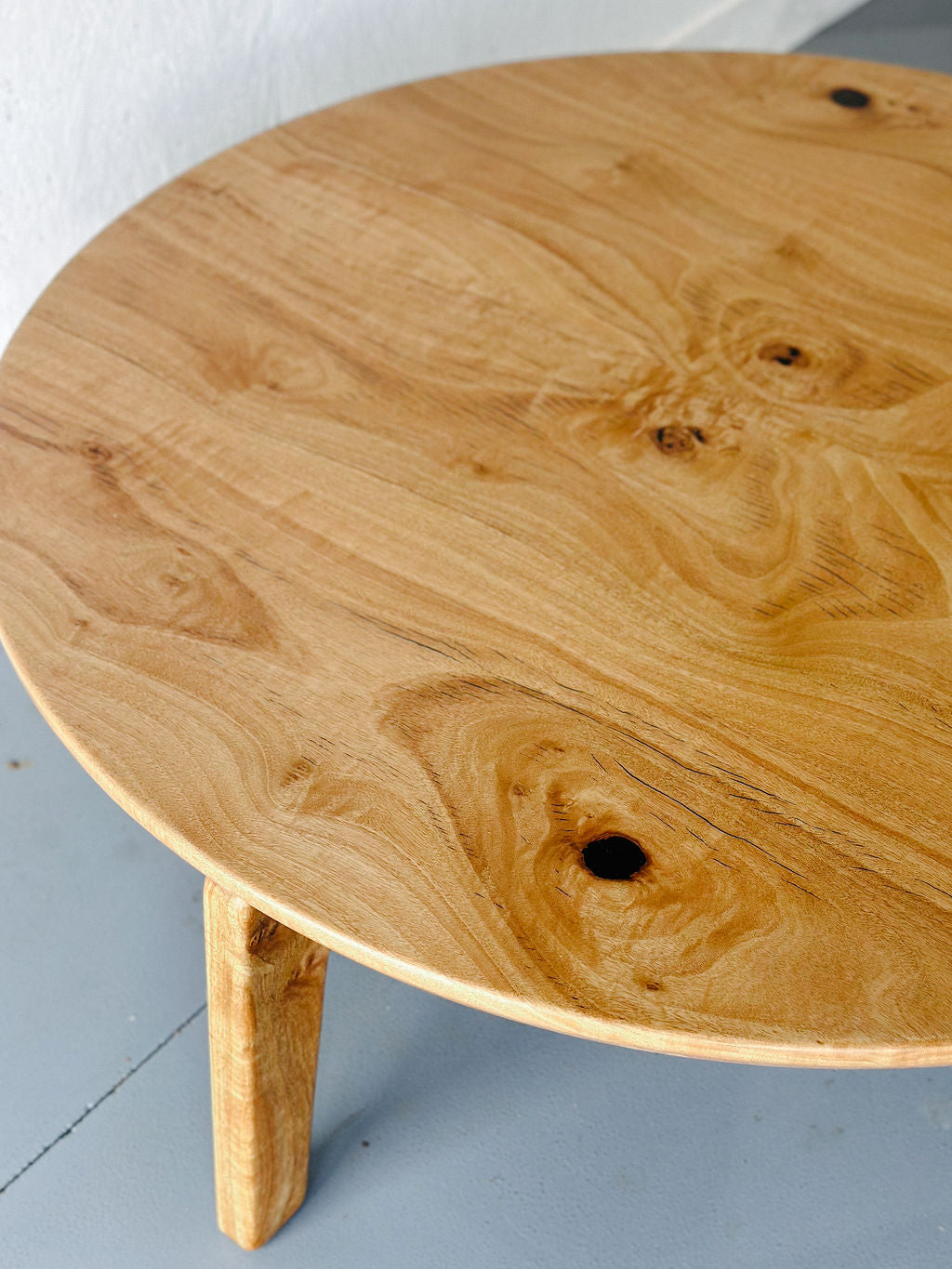 A handcrafted Marri Coffee Table from Timber Grooves with visible knots and natural wood grain patterns, standing on three legs, crafted from exquisite Australian Hardwood, placed on a grey floor.