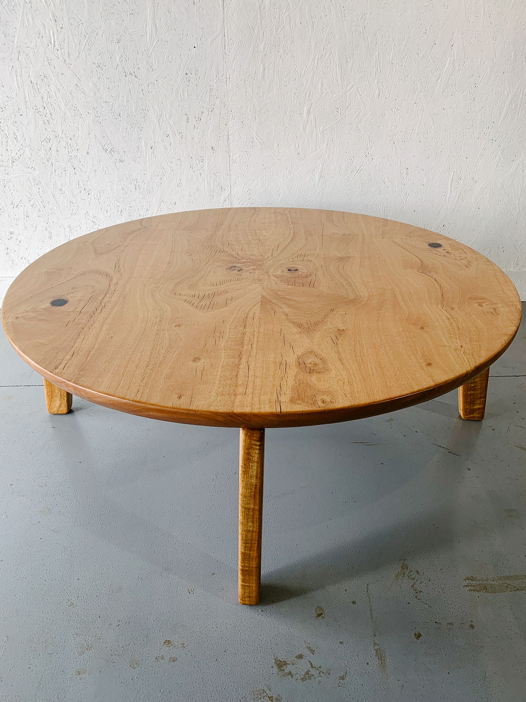 A round Marri Coffee Table with three legs stands on a gray floor, set against a white textured wall. Crafted from Australian hardwood, this handcrafted piece by Timber Grooves adds rustic charm to any space.