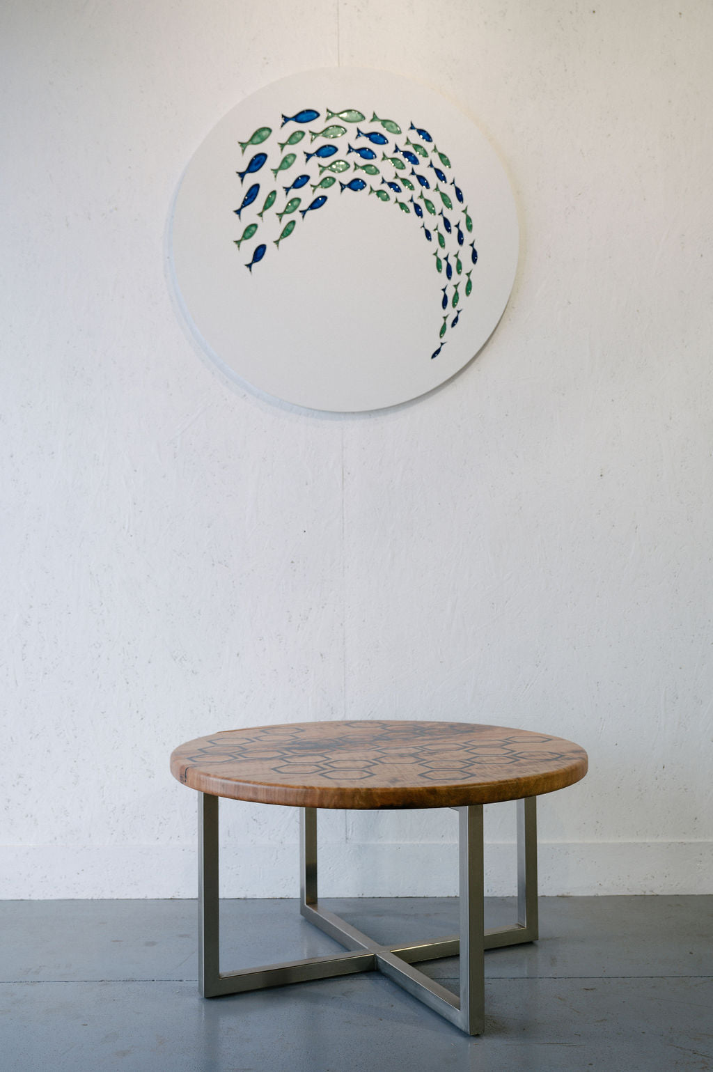 A round wooden table with metal legs is placed against a white wall, with a circular artwork of small, multicolored fish arranged in a wave pattern hanging above it.