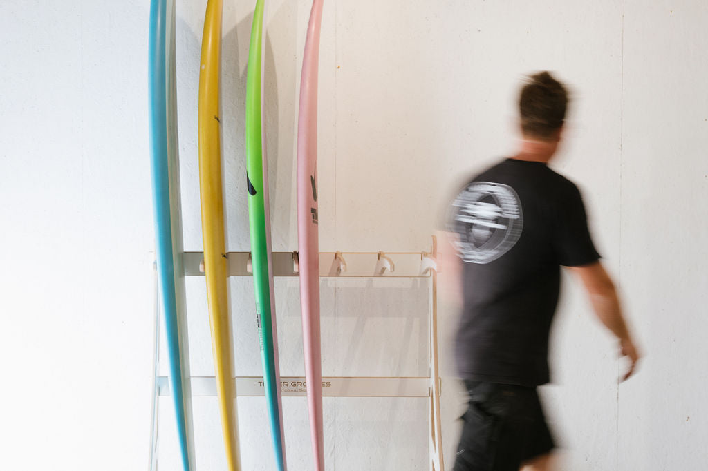 A person in motion walks past a rack holding four surfboards of different colors: blue, yellow, pink, and white.