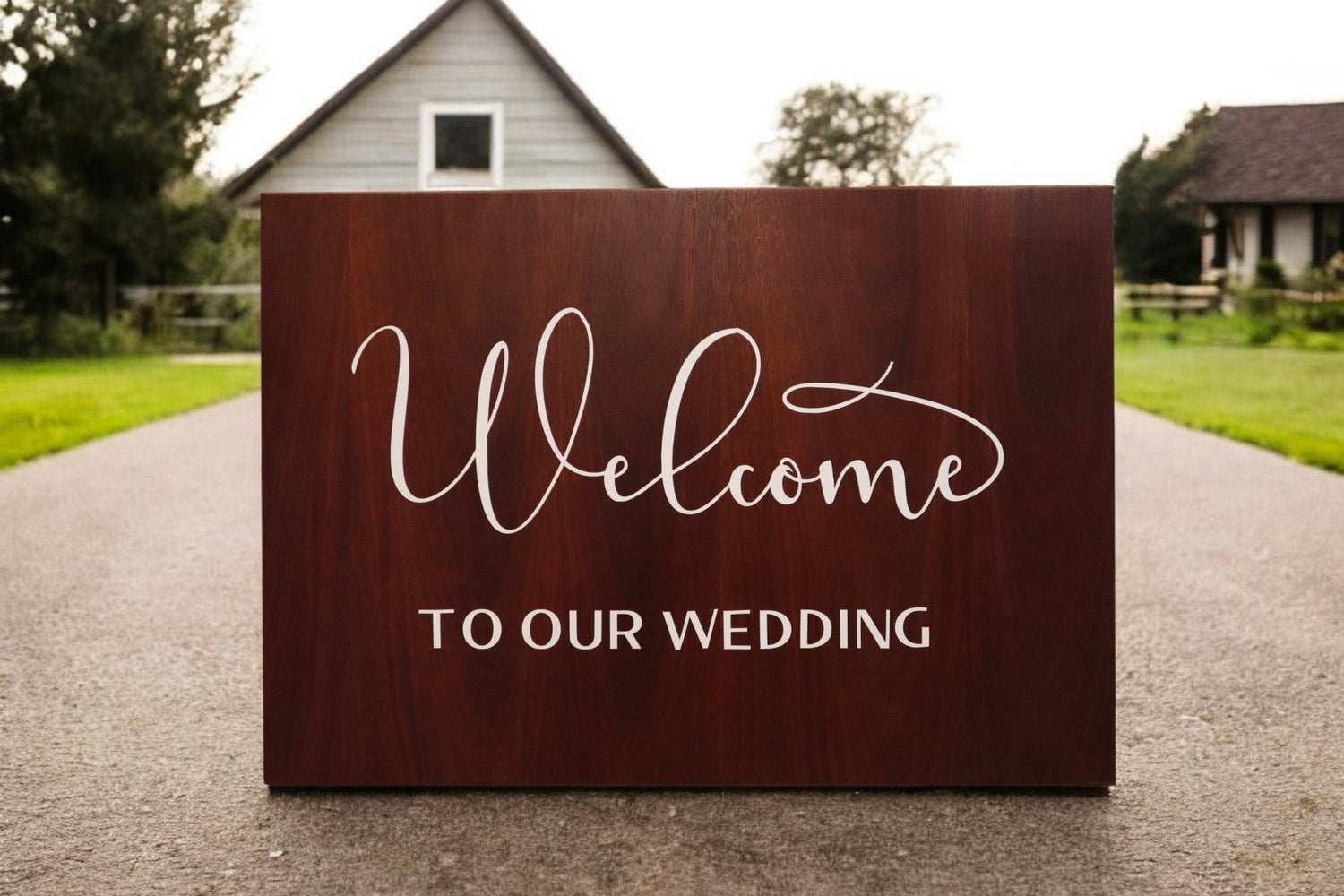 Wooden sign on pathway reads "Welcome to Our Wedding" in elegant script, with houses and greenery in the background.