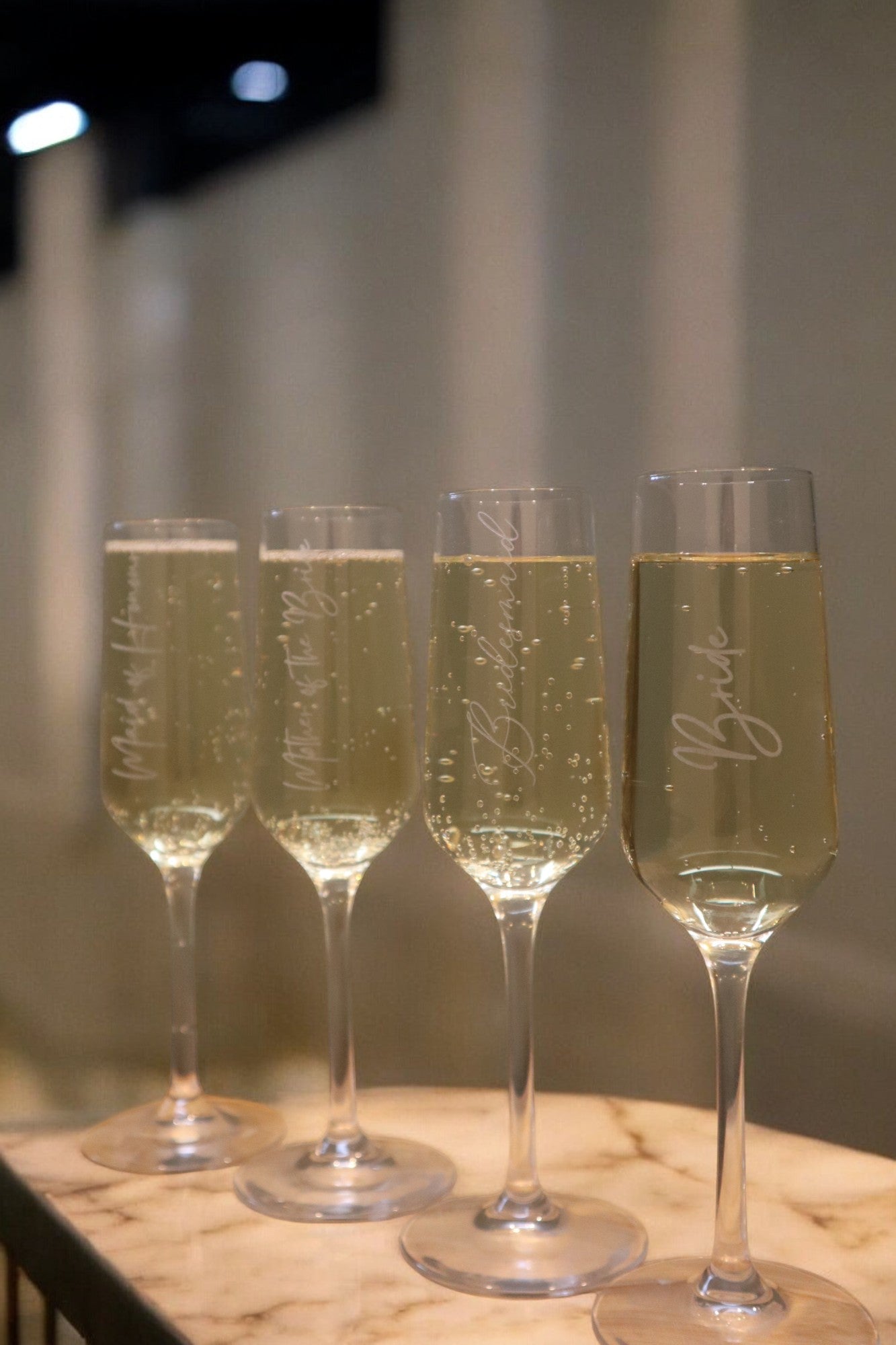 Four champagne flutes filled with bubbly liquid, placed on a marble surface. Each glass has a different engraved inscription.