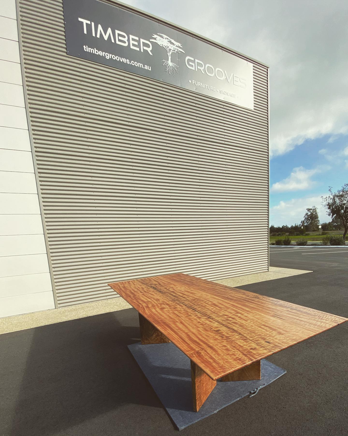 A handcrafted Timber Grooves Marri Dining Table is placed outside a building with the sign "Timber Grooves" on it. The sky is partly cloudy, enhancing the timeless appeal and heirloom-quality of this elegant piece.