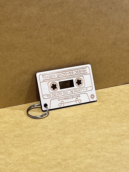 A custom, environmentally conscious wooden keychain resembling a cassette tape, engraved with "Save The Date Casette Keyring" by Timber Grooves and "To Death Do Us Party III.