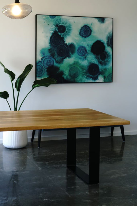 An American White Oak Dining Table from Timber Grooves with a black steel base stands on a polished concrete floor. Behind it is a potted plant and wall art featuring blue and green abstract patterns. A ceiling light hangs above, illuminating the handcrafted dining room furniture.