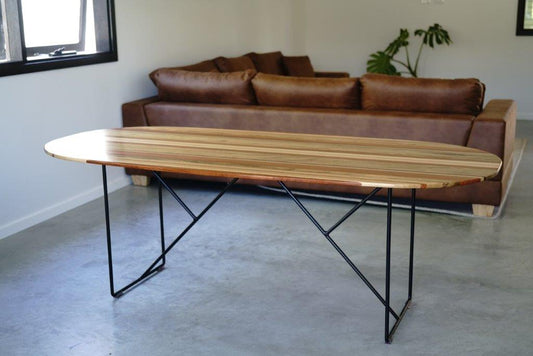 A Timber Grooves Surfboard Dining Table with unique grain patterns and black metal legs is positioned on a concrete floor in front of a brown leather sofa in a minimalist living room. A potted plant is in the background.