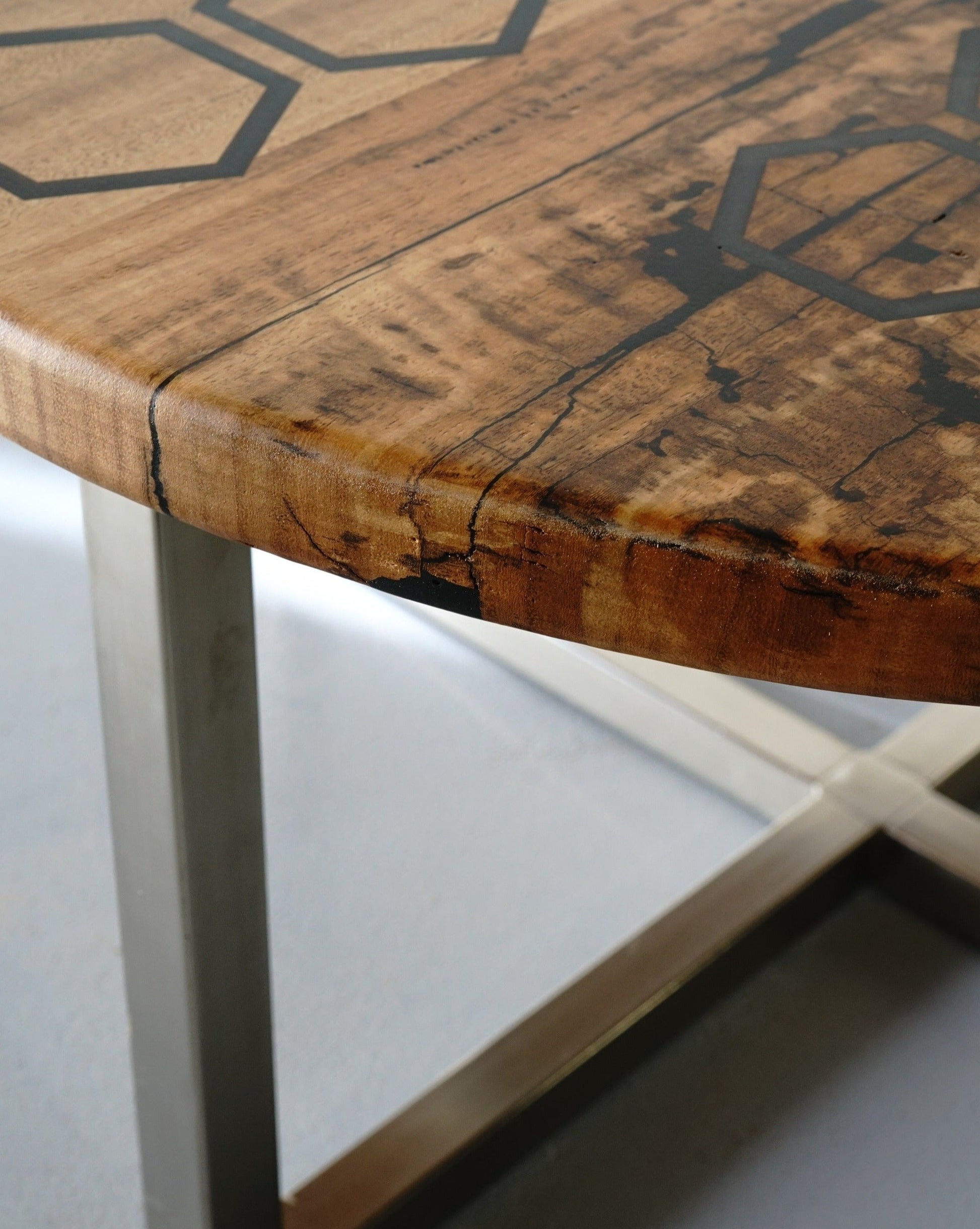 Close-up of the Marri Coffee Table by Timber Grooves, with a stainless steel base and hexagon patterns inlaid on the tabletop. The wood features natural cracks and visible grain texture, complemented by a black resin inlay for a touch of modern design.