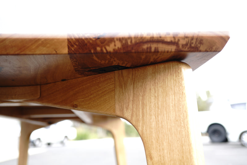 Close-up view of the handcrafted Marri Dining Table leg and edge by Timber Grooves, showing the exquisite grain and texture of the wood. The background is blurred, with a white car and greenery visible.