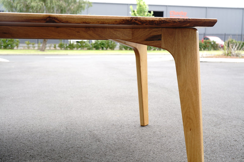 Close-up of a handcrafted Marri Dining Table by Timber Grooves with sleek, angled legs, set on an outdoor asphalt surface. A building with a red sign is visible in the background.