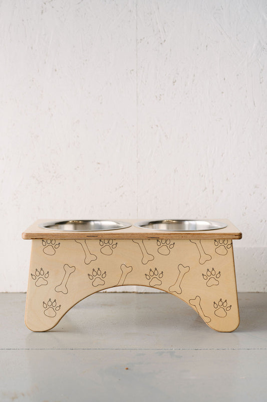 A Timber Grooves Double Raised Dog Bowl Stand made of wood with carved paw prints and bone designs, featuring two stainless steel bowls to help reduce symptoms of bloat and alleviate skeletal and joint pain.