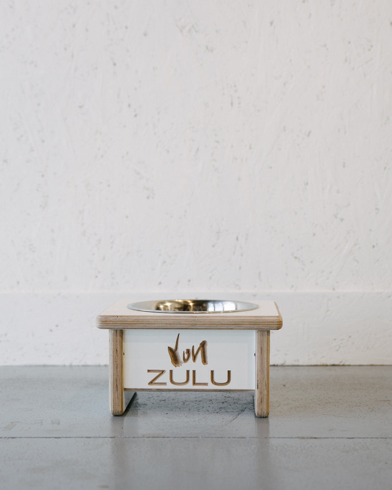 A pet food bowl with a wooden dog bowl stand labeled "Dog Bowl Stand Custom Engraving" by Timber Grooves sits on a gray floor against a textured white wall, featuring custom engraving based on customer notes.