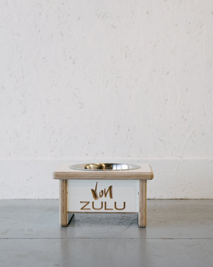 A Large Timber Grooves Single Raised Dog Bowl Stand with stainless steel bowls and custom engraving of "Von Zulu" on the front, placed against a textured white wall.