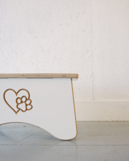 White wooden bench with a heart and paw print cutout design on its side, placed on a light grey floor with a textured white wall in the background. Ideal for pet lovers, it pairs beautifully with the Timber Grooves Dog Bowl Stand Custom Engraving to create a cozy corner.