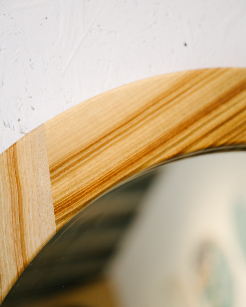 Close-up view of a Timber Grooves Camphor Laurel Mirror with a smooth, light wood finish against a white textured wall.