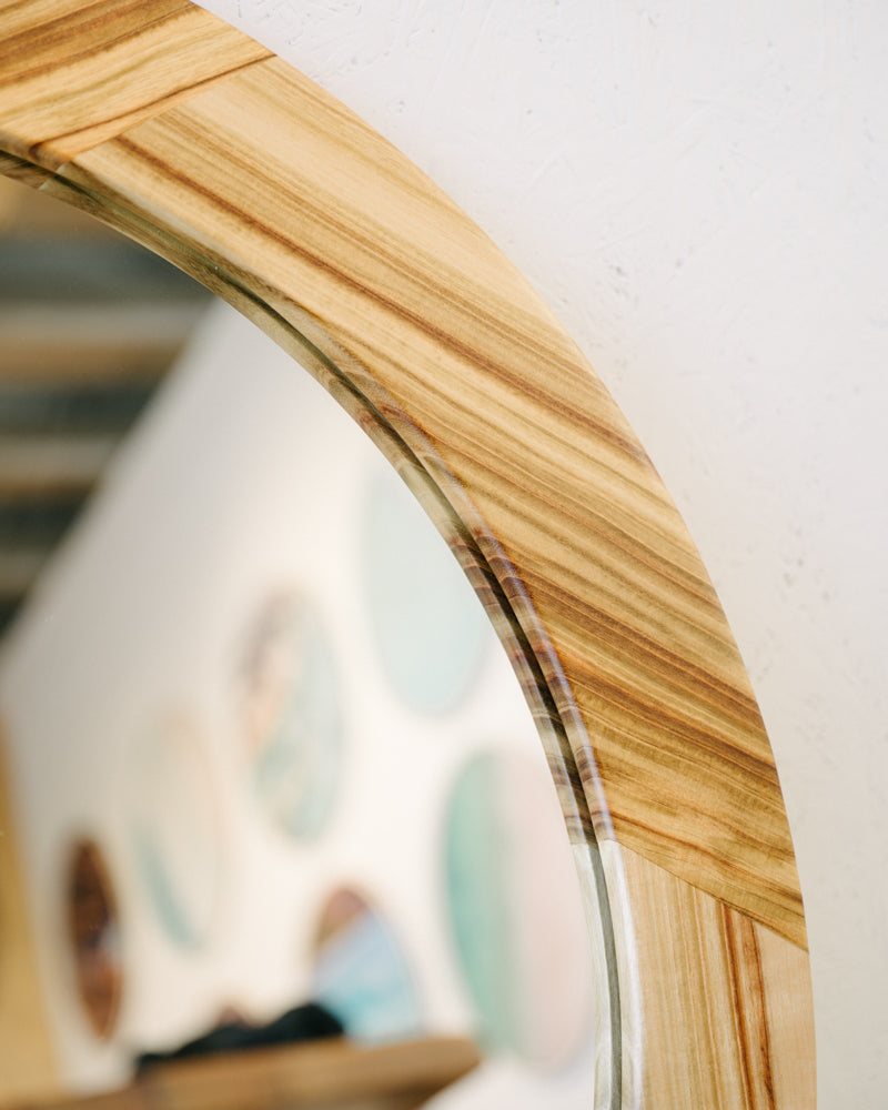 Close-up of a unique circular frame made from camphor laurel, the Camphor Laurel Mirror by Timber Grooves, reflecting a blurred wall adorned with round, colorful artworks.