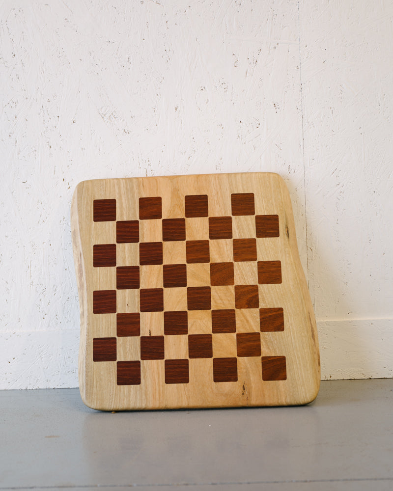 A Timber Grooves Marri Chessboard with Jarrah Inlay with a checkered pattern composed of alternating light and dark wood squares, positioned against a white textured wall.