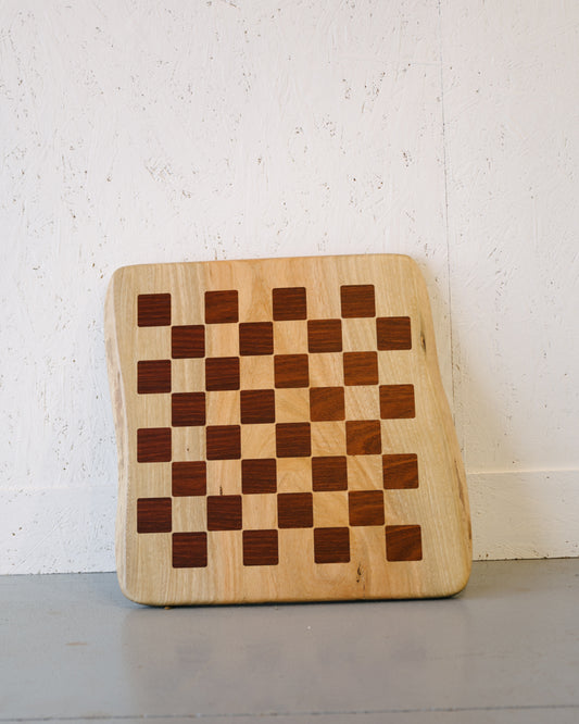 A Timber Grooves Marri Chessboard with Jarrah Inlay with a checkered pattern composed of alternating light and dark wood squares, positioned against a white textured wall.