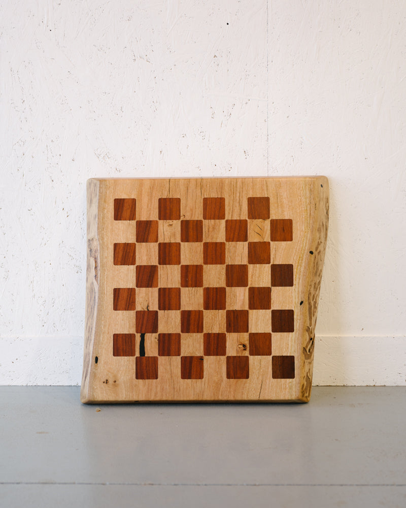 A rectangular wooden cutting board, reminiscent of a Timber Grooves Marri Chessboard with Jarrah Inlay with its checkered pattern of light and dark wood squares, rests on a light gray floor against a white textured wall.