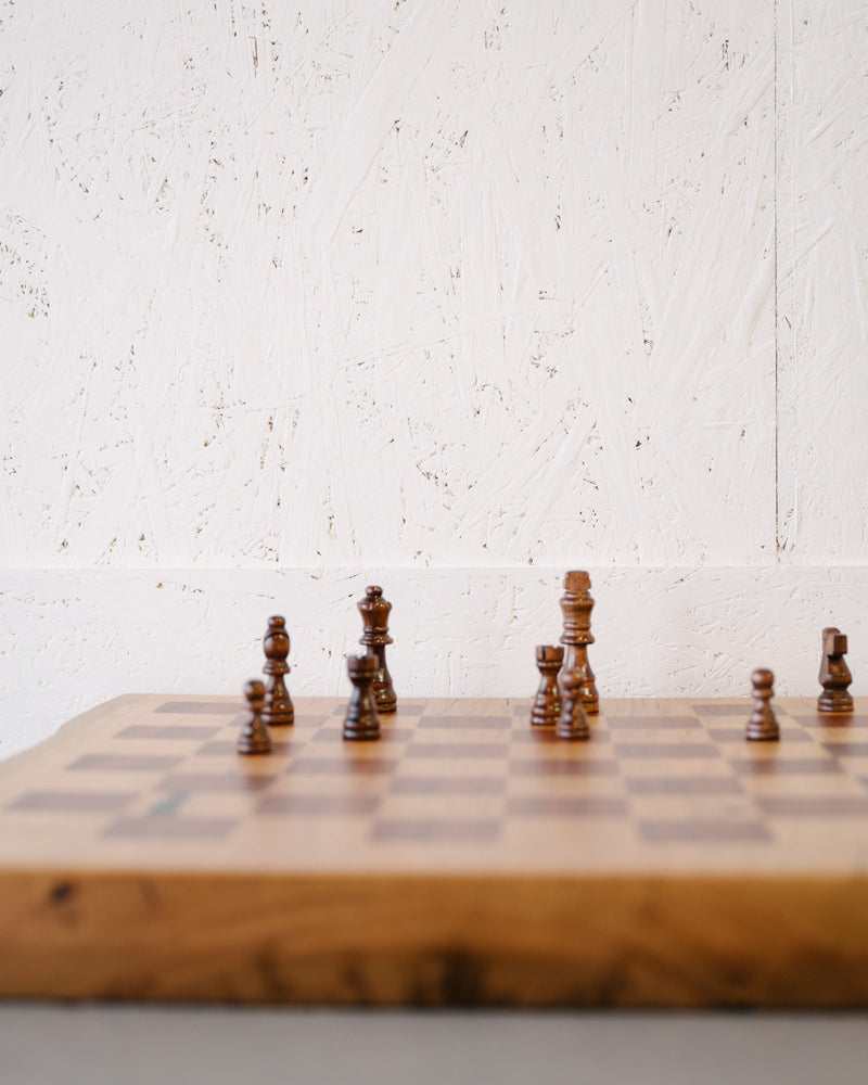 A Marri Chessboard with Jarrah Inlay by Timber Grooves in mid-game setup, positioned on a premium marri slab table against a textured white wall background. Perfect for intense chess games.