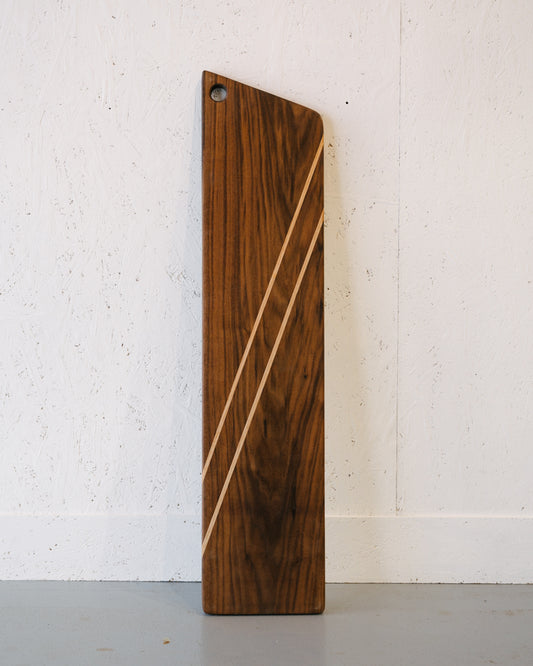 Vertical wooden cutting board with diagonal light-colored American Oak inlay stripes, set against a white textured background and featuring a small hole near the top edge. This handcrafted piece, the Timber Grooves Walnut Charcuterie Board, exudes elegance and functionality.