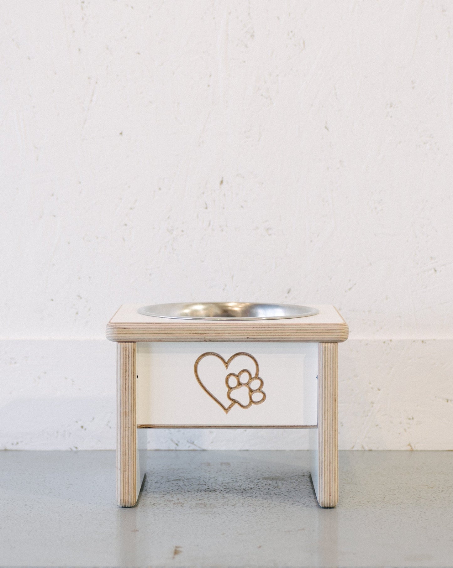 A Large Raised Single Dog Bowl Stand with custom engraving, featuring a heart and paw print design, is placed on a light-colored floor against a white wall.