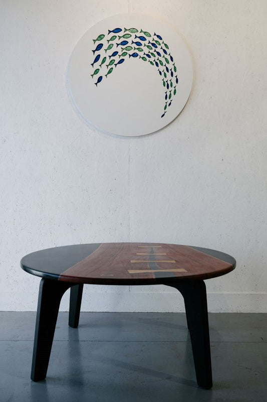 A Timber Grooves Jarrah Tuxedo Coffee Table with a dark surface and wood accents is positioned in front of a circular wall art piece depicting a fish design.