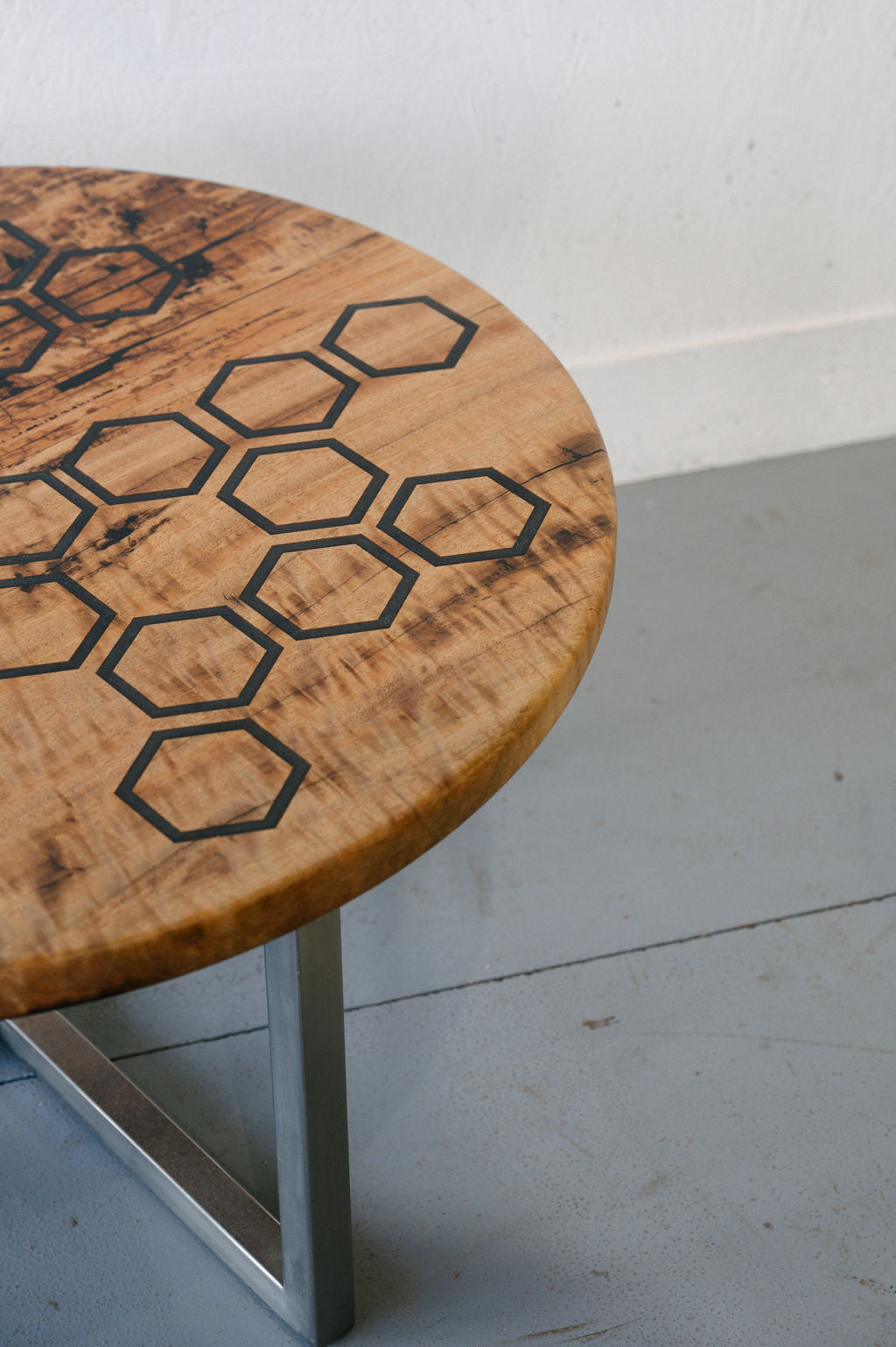 A round wooden Marri Coffee Table by Timber Grooves with a geometric hexagonal design, black resin inlay, and metal legs, placed on a concrete floor.